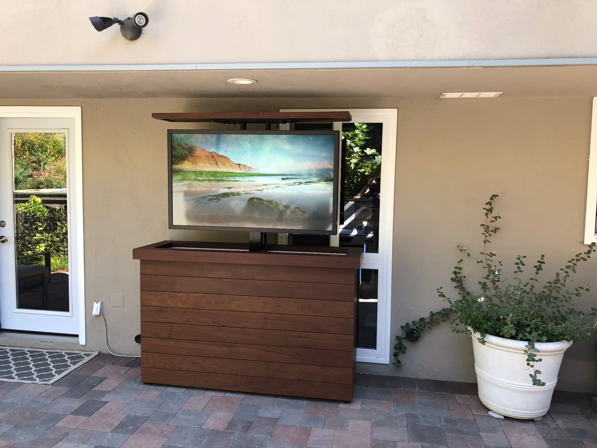 Motorized outdoor TV lift cabinet in warm wood finish rising from patio entertaining area