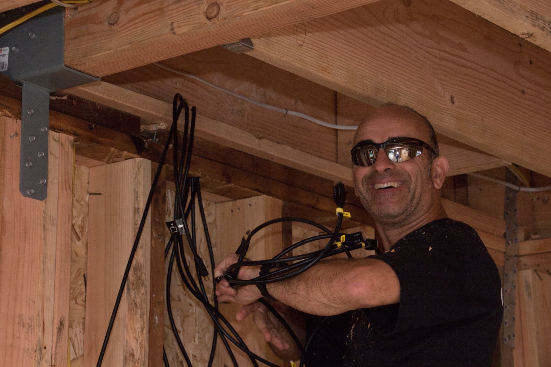 Low-voltage technician smiling while routing a bundle of black cables through unfinished framing