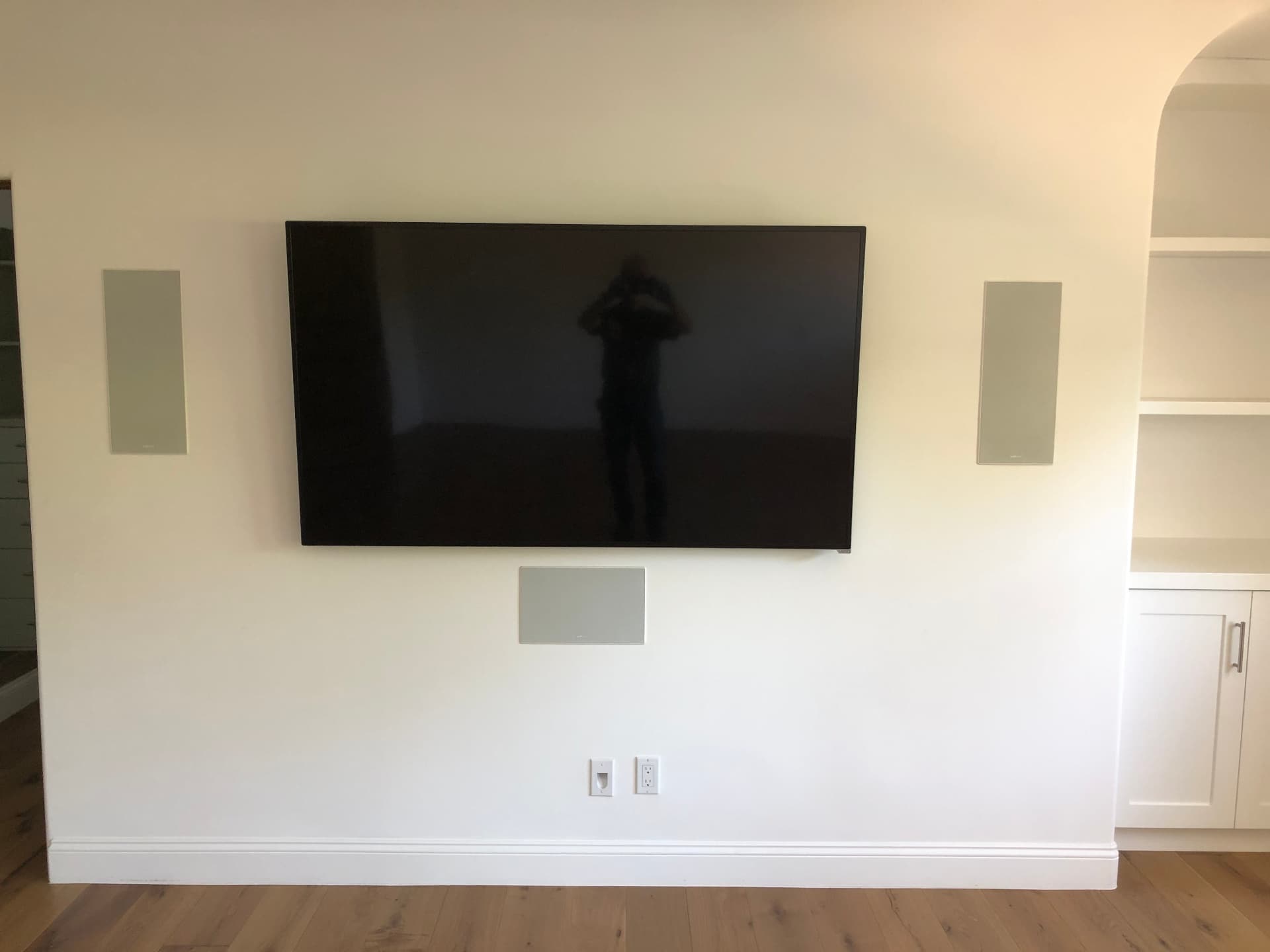 Flat-screen TV flush-mounted on white wall with in-wall left, right, and center channel speakers in modern living room with hardwood floors